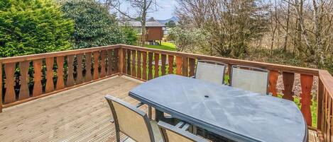 Una terraza con mesa y sillas, rodeada por una baranda de madera, con árboles y una casa al fondo.