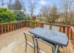 A deck with a table and chairs, surrounded by a wooden railing, with trees and a house in the background.