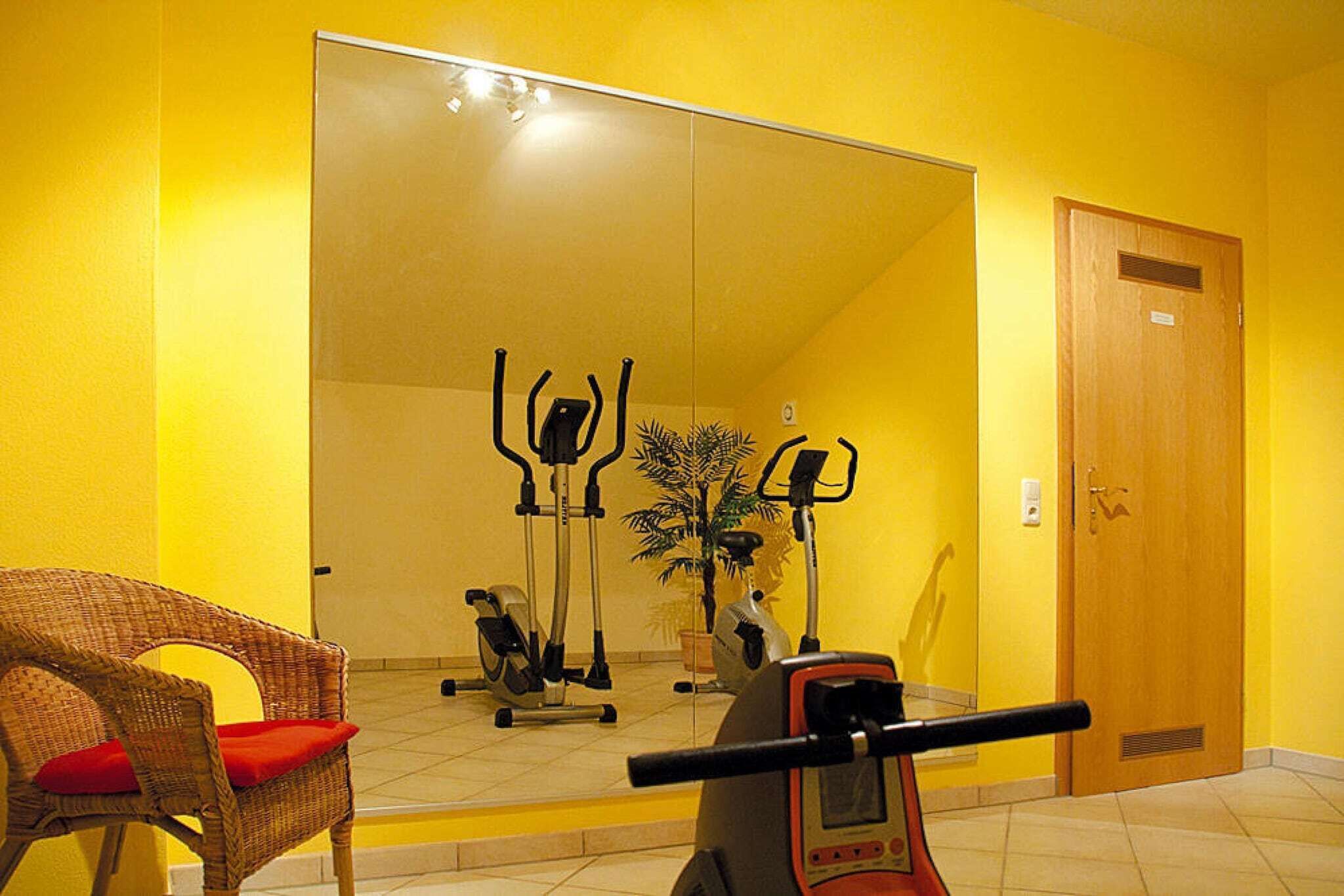 Una habitación con un espejo grande que refleja equipo de ejercicio, una silla de mimbre con cojín rojo y una puerta de madera.