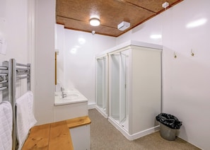 Une salle de bain avec un banc en bois, deux lavabos, une cabine de douche en verre, une poubelle blanche et un luminaire au plafond.