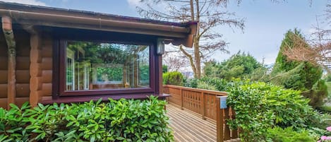 Una cabaña de troncos con un ventanal grande, una terraza de madera y abundante vegetación.
