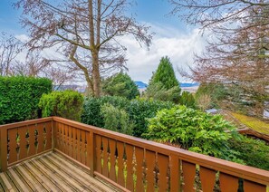 A wooden deck with a railing, surrounded by various green shrubs and trees.