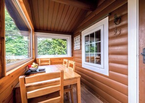 Una cabaña con interior de madera, una mesa de comedor con sillas y dos ventanas con vistas al follaje.