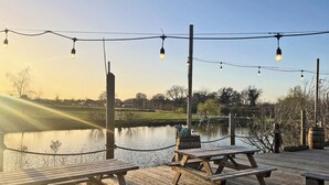 Een houten terras met picknicktafels met uitzicht op een rustig wateroppervlak, sfeerverlichting en een heldere lucht.