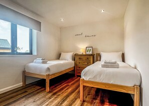 Un dormitorio con dos camas individuales, un aparador, una lámpara de noche, un cuadro enmarcado y una ventana con vistas.