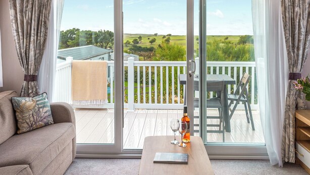 A room with a sofa, a table with a bottle and glasses, and a view of a deck and landscape through large sliding doors. - 3 bedroom accommodation in Holywell Bay (Holywell Bay)