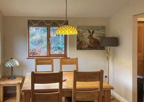 Un coin repas avec une table et des chaises en bois, une fenêtre donnant sur un paysage verdoyant, un luminaire suspendu jaune et un tableau représentant un lapin accroché au mur.