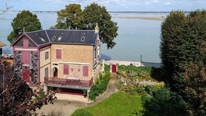 Ein zweistöckiges Haus mit roter Tür und einem Balkon, umgeben von einem gepflegten Garten und Blick auf ruhiges Gewässer.