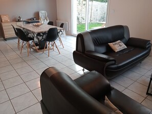 A living room with a black leather sofa, a dining table with chairs, and a view of the outdoors. - Gite Proche Baie de Somme (Arrest)