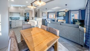 Una cocina moderna con electrodomésticos integrados, una zona de comedor con una mesa de madera y bancos acolchados, y una sala de estar con sofá y televisor.