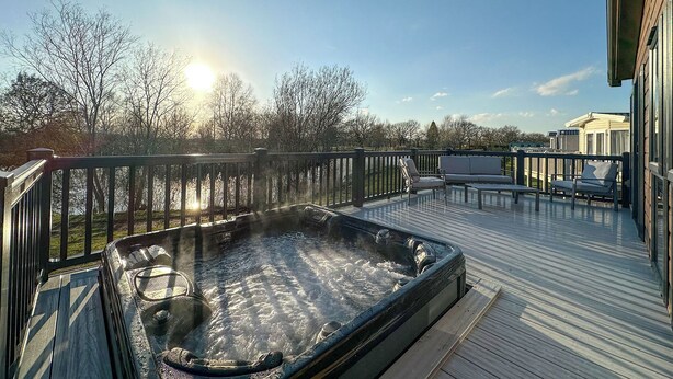Un jacuzzi en una terraza de madera con vista a un lago y árboles, y un edificio al fondo.