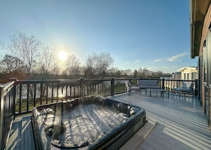 Un jacuzzi en una terraza de madera con vista a un lago y árboles, y un edificio al fondo.