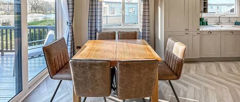 A dining area with a wooden table and cushioned chairs, a kitchen with cabinets, and a view through large windows.