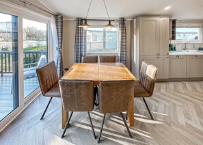 A dining area with a wooden table and cushioned chairs, a kitchen with cabinets, and a view through large windows.