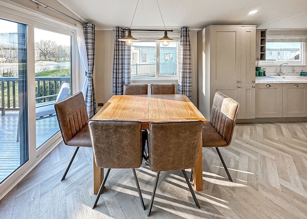 Un comedor con mesa de madera y sillas acolchadas, una cocina con armarios, y vistas a través de amplios ventanales.