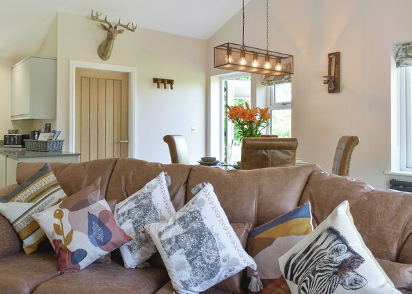A living room with a brown leather sofa, patterned cushions, a deer head mounted on the wall, a dining area with a table and chairs, and a kitchen in the background.