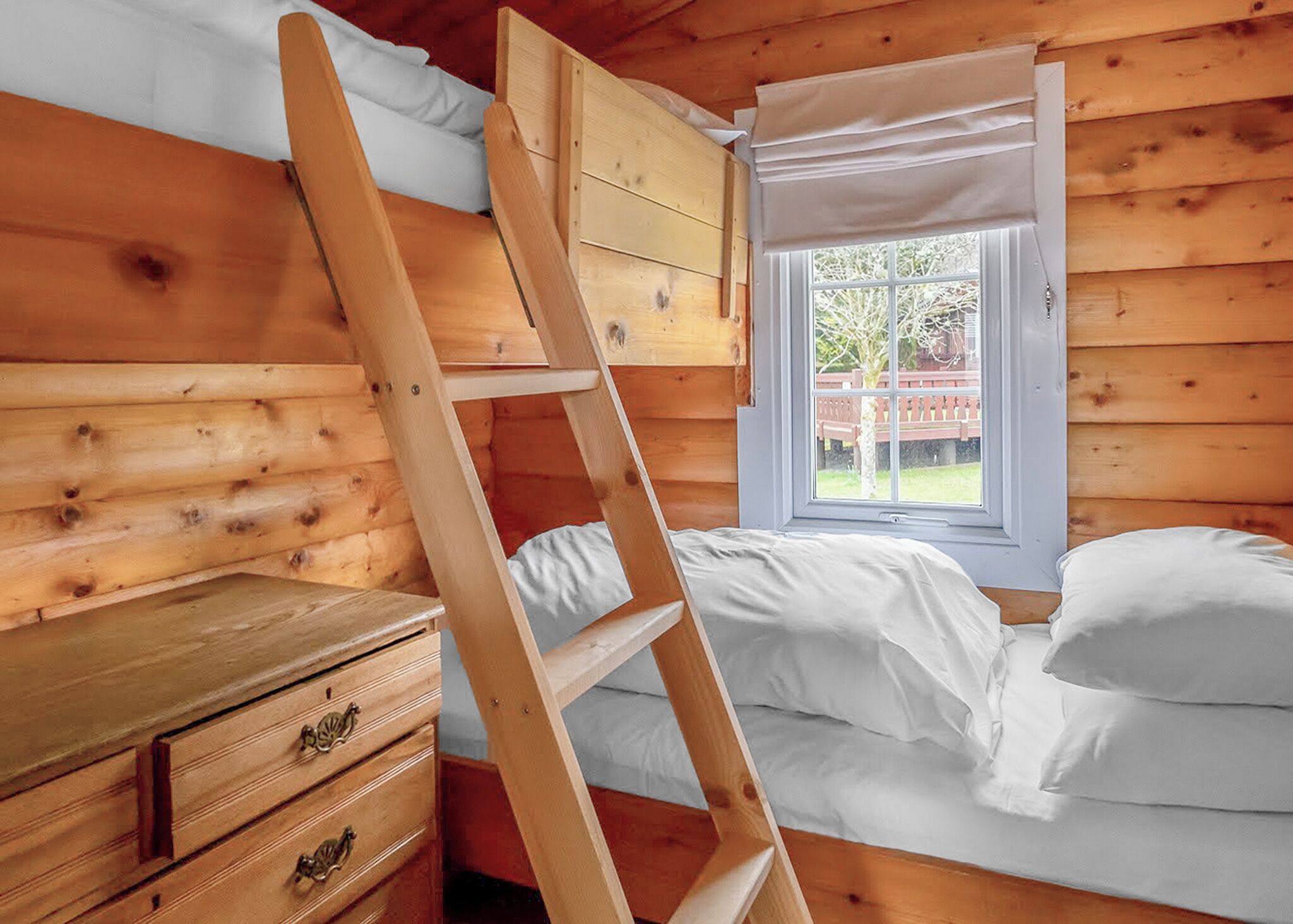 Ein Holzhütten-Zimmer mit einem Etagenbett, einem Fenster mit Blick auf einen Zaun und einer Holzkommode mit Schubladen.