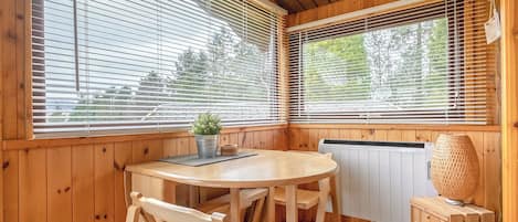 Interior de una cabaña de madera con una mesa redonda, dos sillas, una planta en maceta, un radiador y una lámpara de fibras naturales.