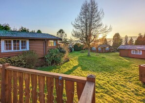 A log cabin with a wooden deck, surrounded by a grassy yard and a large tree.
