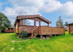 A wooden cabin with a deck, surrounded by a grassy yard and trees.