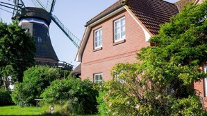 A traditional brick house with a windmill in the background, surrounded by lush greenery. - Kuschelige Küstenlodge Lichtmeer (Nordstrand)