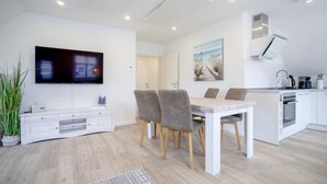 A modern living room with a flat-screen TV, a dining area with a table and chairs, a kitchen with white cabinets, and a large window. - Kuschelige Küstenlodge Lichtmeer (Nordstrand)