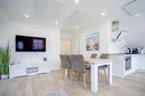 Een moderne woonkamer met een flatscreen-tv, een eethoek met een tafel en stoelen, een keuken met witte kasten, en een groot raam.