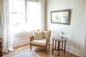 Una habitación con una silla de madera, una pequeña mesa redonda con un florero y un cuadro en la pared.