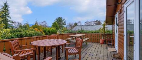 A wooden deck with a table and chairs, a glass door, and a view of a garden.