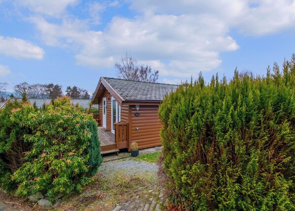 A small wooden cabin with a door and windows, surrounded by dense greenery and a gravel path.