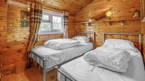 A wooden cabin room with two metal beds, a window with curtains, and a hanging light fixture.