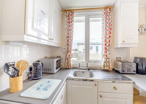 Una cocina con armarios blancos, un lavamanos, dos tostadoras, un hervidor y una ventana con cortinas estampadas.