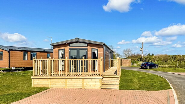 A wooden mobile home with a balcony, a paved driveway, and a car parked nearby. - 1 bedroom accommodation in Tunstall, Nr Hull (Tunstall, Nr Hull)