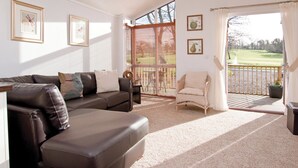A spacious living room with a brown leather sofa, a wicker chair, and a large window overlooking a golf course. - 1 bedroom accommodation in Narberth, Nr Tenby (Narberth, Nr Tenby)