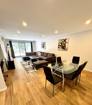 Een moderne woonkamer met een glazen eettafel, zwarte stoelen, een bruine bank en een zwarte vloerlamp.