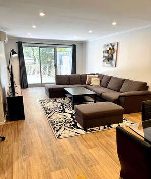 Een moderne woonkamer met een hoekbank, een salontafel en een glazen eettafel. Er is een groot raam met gordijnen, een zwarte vloerlamp en een schilderij aan de muur.