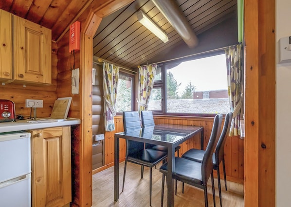 A cabin interior with a wooden interior, a dining table with chairs, a refrigerator, and a window with curtains.