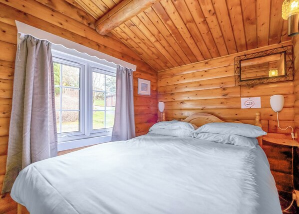 A wooden cabin bedroom with a bed, curtains, a window, a bedside table, and a wall-mounted light fixture.