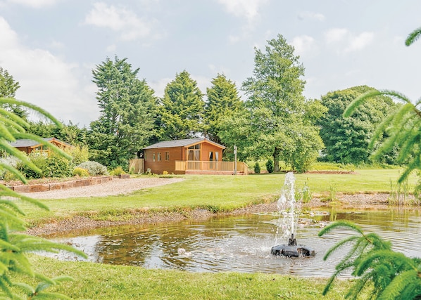 A wooden cabin with a deck, surrounded by a pond and lush greenery. - 2 bedroom accommodation in Narberth, Nr Tenby (Narberth, Nr Tenby)