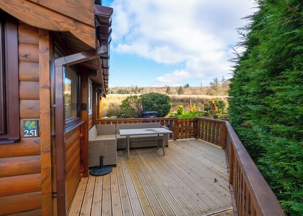 Una terraza de madera con una mesa y sillas, un letrero de número y vistas a un paisaje cubierto de hierba.