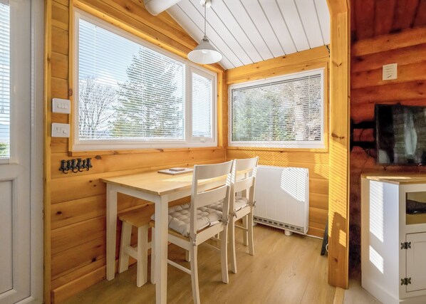 A wooden cabin interior with a dining table, chairs, a white radiator, and a window with blinds.