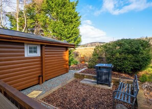 A log cabin with a small window, a gravel patio, a wooden bench, and a garden area with mulch and plants.