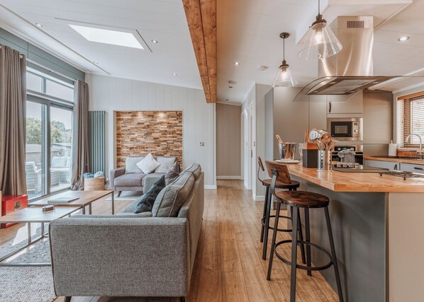 Una sala moderna con un sofá, un comedor con taburetes de bar, una cocina con campana de acero inoxidable y un ventanal que proporciona luz natural.