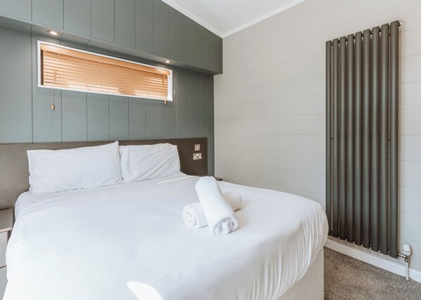 A neatly made bed with white linens and a rolled towel, a headboard, a window with blinds, and a radiator.