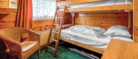 Habitación tipo cabaña de madera con literas, una silla de mimbre, una escalera y una ventana con cortinas.