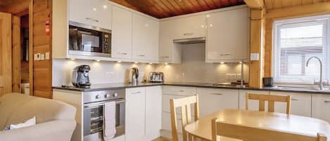 Cozinha compacta com armários brancos, forno embutido, cafeteira e área de jantar com mesa e cadeiras de madeira.