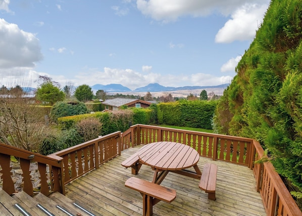Une terrasse en bois avec une table de pique-nique et des bancs, entourée de verdure et offrant une vue sur les montagnes lointaines.