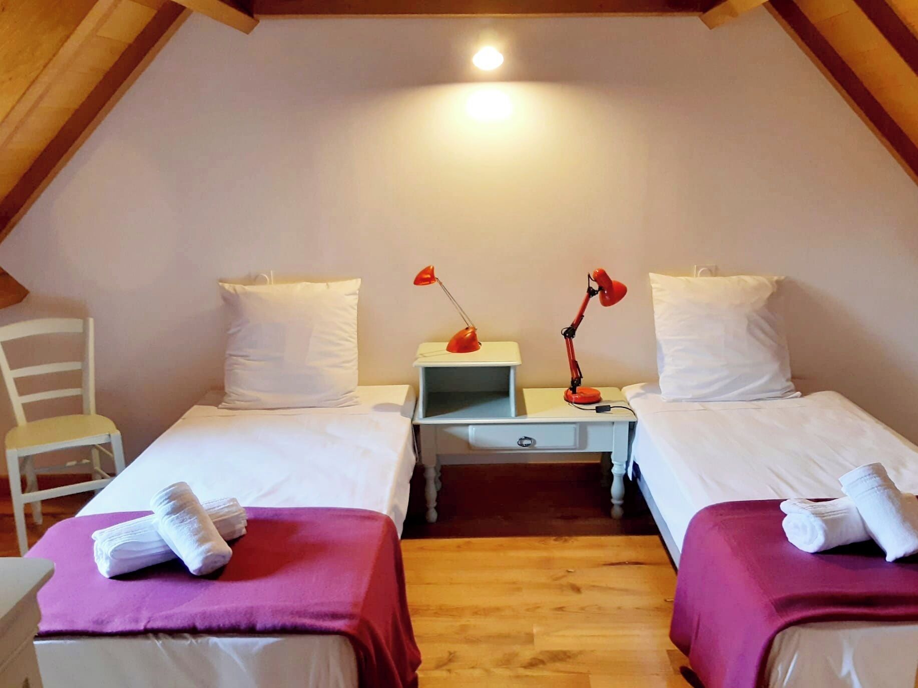 Une chambre avec deux lits, chacun étant équipé d’un oreiller et d’une serviette enroulée. On y trouve deux tables de chevet avec des lampes, et une chaise est visible dans un coin.