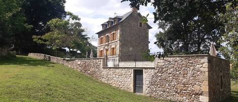 A stone building with a balcony and a door, surrounded by a stone wall and a grassy area.