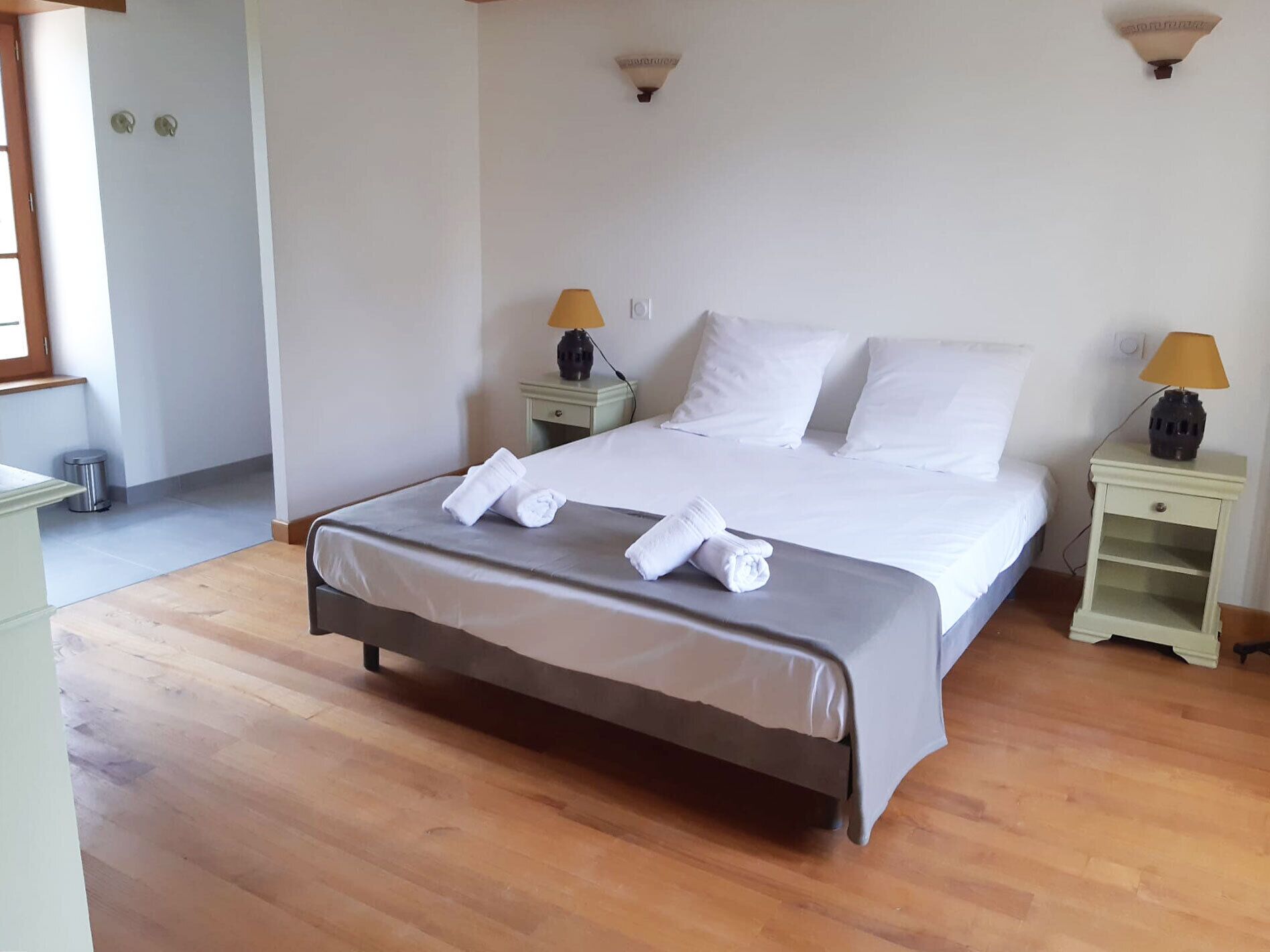 Un letto rifatto con lenzuola e asciugamani bianchi, due comodini con lampade e un pavimento in legno.